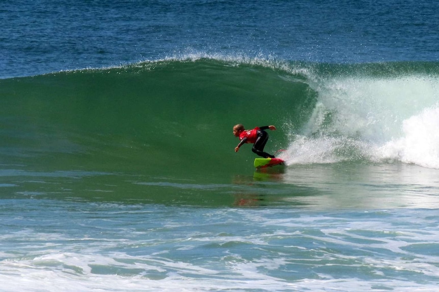 A young girl wearing a wetsuit and red rash vest surfs a wave.