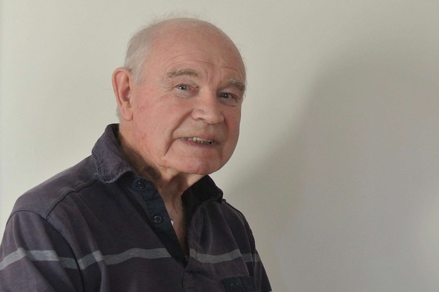 A balding old man wearing a blue shirt with blue stripes smiles at the camera.