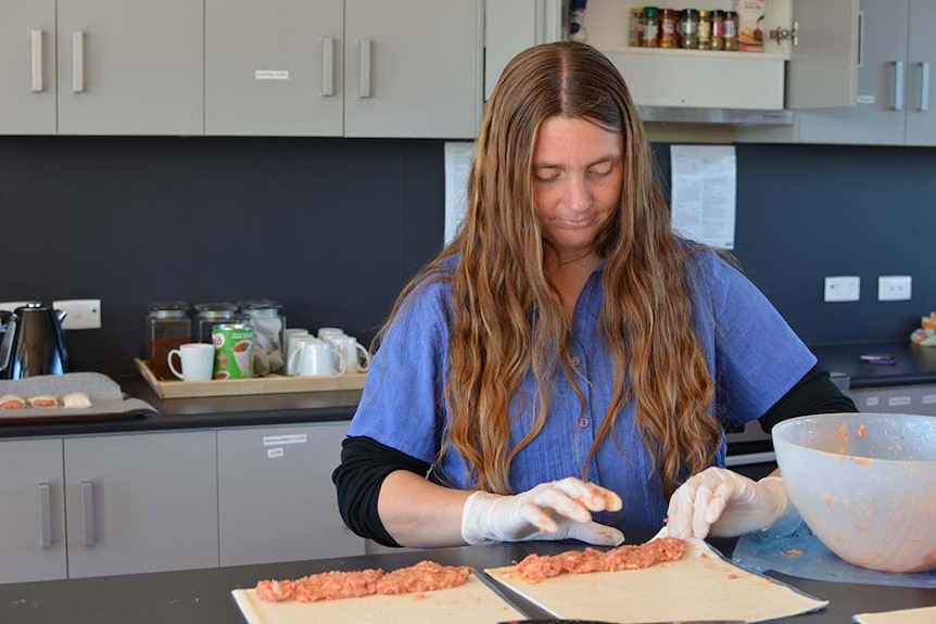A woman with long dark blonde hair stands in a kitchen rolling mince into pastry to make sausage rolls.