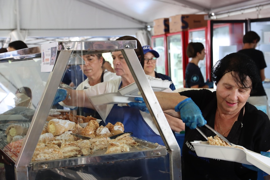 Women wearing hairnets serve food from a warmer. They look happy.