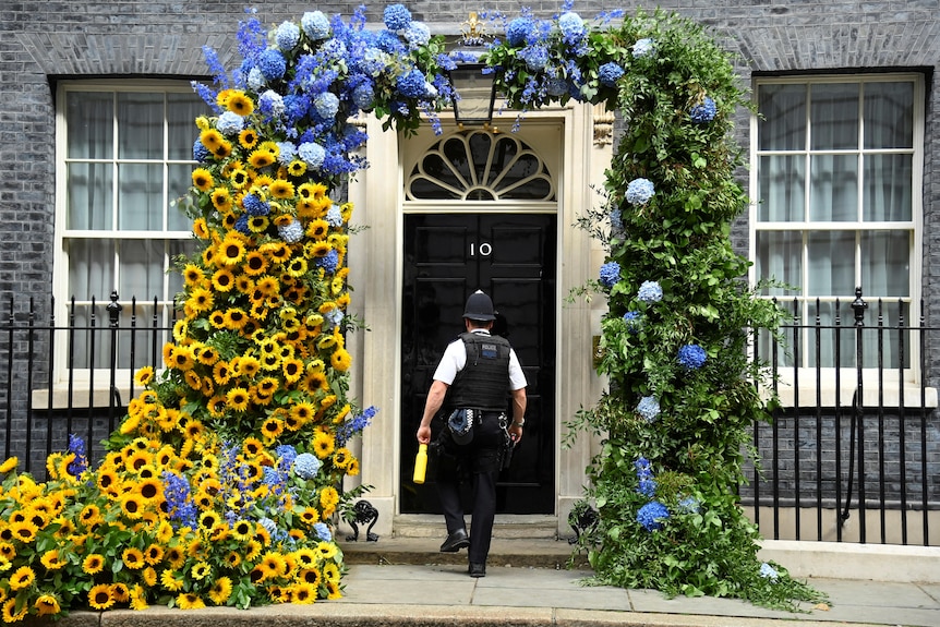 A UK police officer walks through an arch of yellow and blue flowers in front of the door to 10 Downing Street.