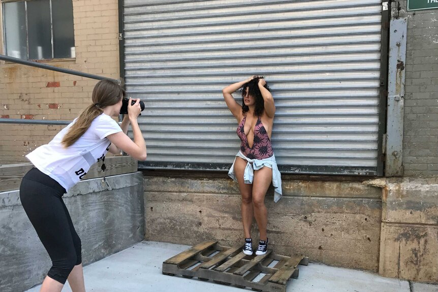 A busty woman in a one-piece swimsuit poses for a female photographer in a studio