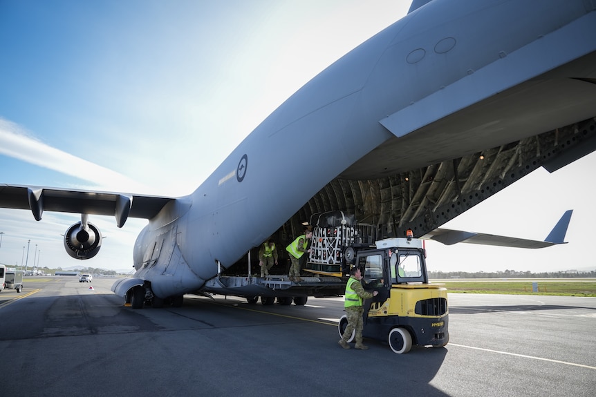 Large, grey military aircraft at airport. Pallets of supplies are loaded by people in army uniforms and high-vis vests.