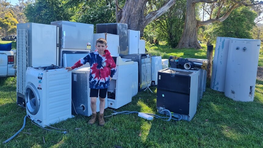 A boy stands in front of a pile of white goods in a back yard.