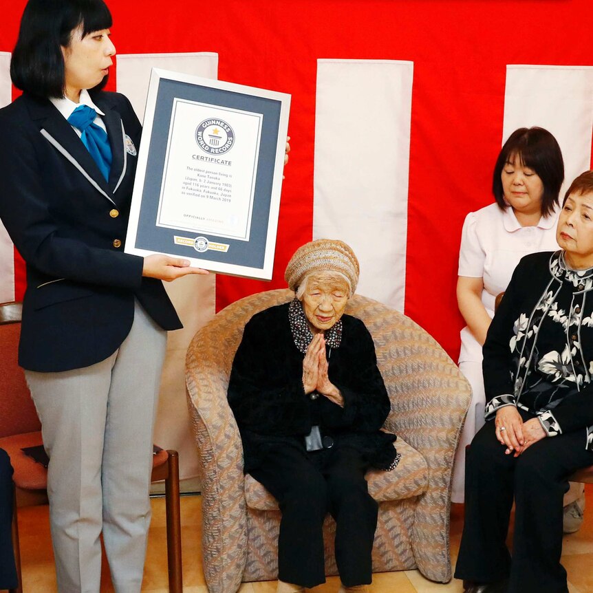 Against a bright red backdrop, three women gather around Kane Tanaka, with one woman in Guinness World Records uniform.