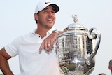 Brooks Koepka smiles while leaning his arm on the Wanamaker Trophy