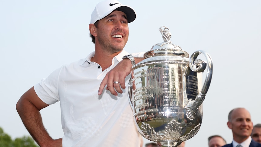 Brooks Koepka smiles while leaning his arm on the Wanamaker Trophy