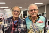 Two smiling elderly men, both wearing glasses,, grey hair, one has handlebar moustache and in a room with other people behind.