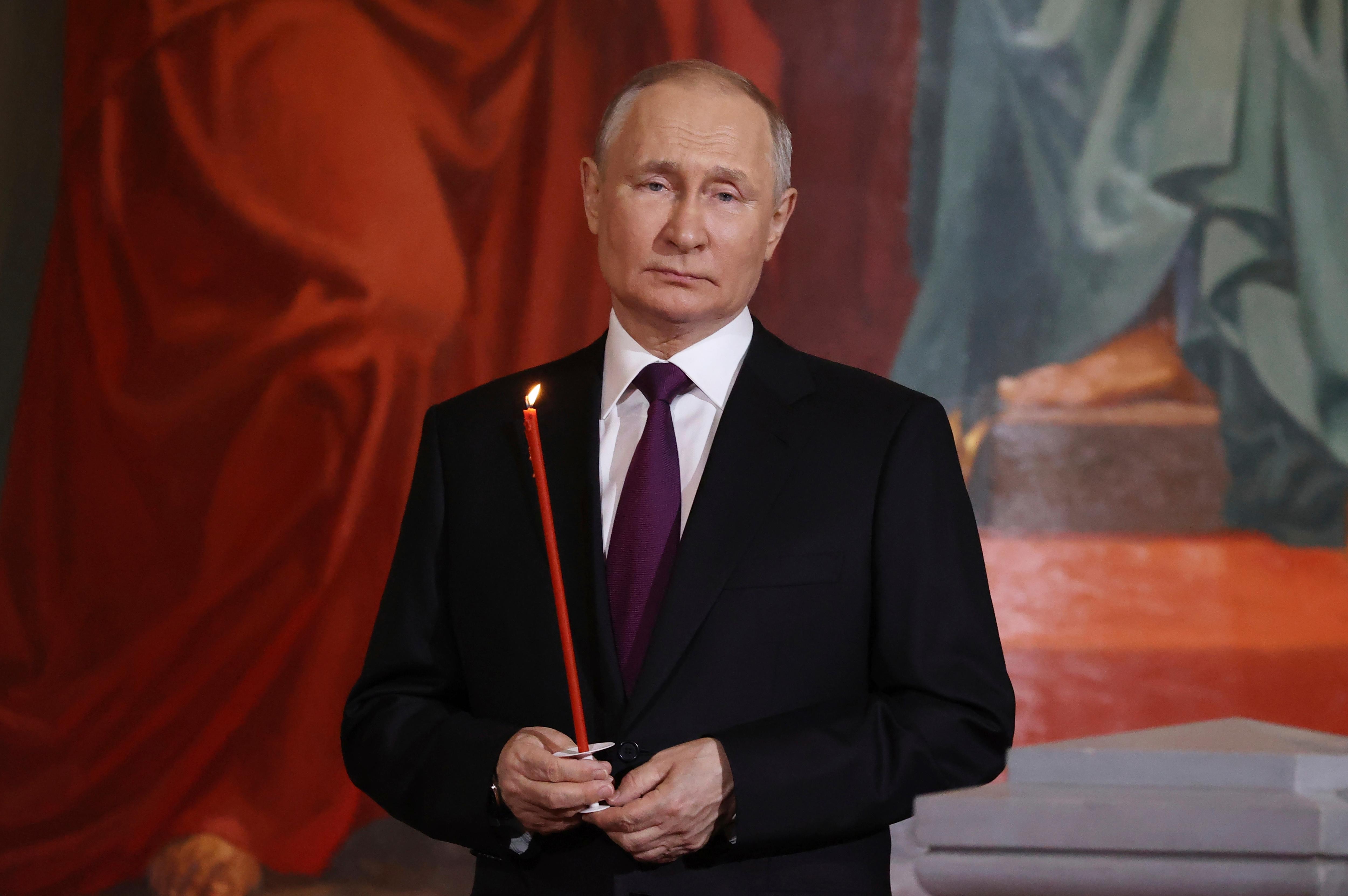 Putin, wearing a suit, holds a lit thin red candle in front of a mural background
