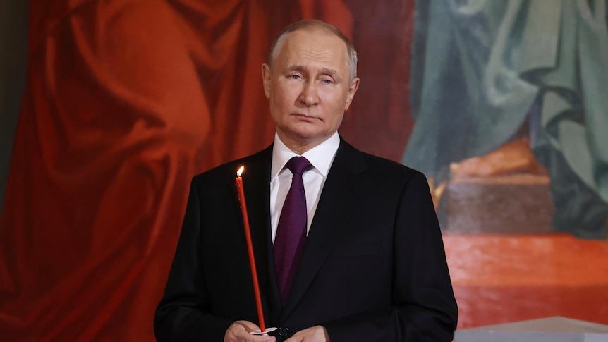 Putin, wearing a suit, holds a lit thin red candle in front of a mural background