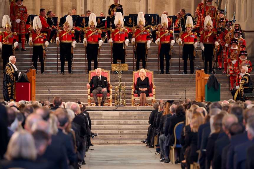 King Charles and Camilla sit in two chairs in the middle of a staircase with a crowd in front and officials behind. 