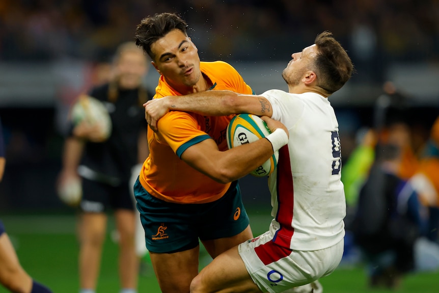 An Australian male rugby international holds the ball as he is tackled by an England opponent.