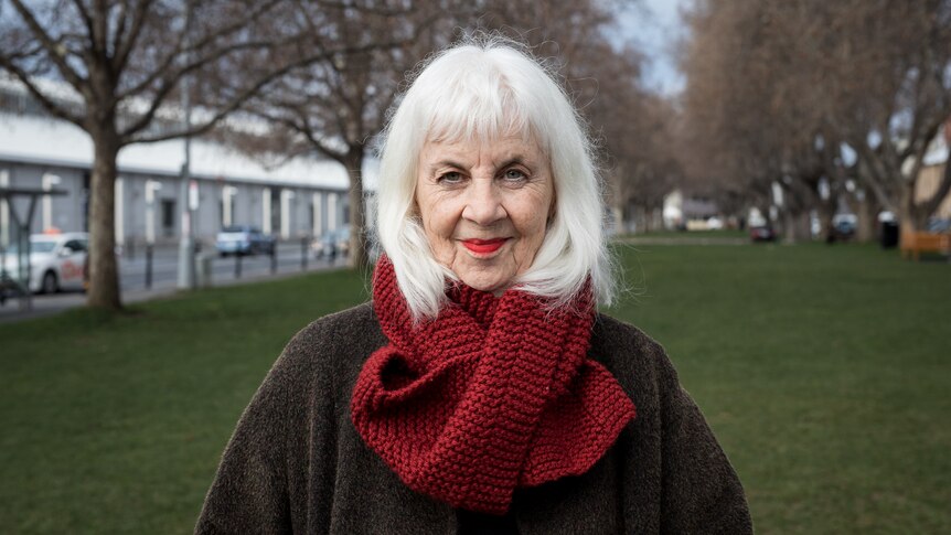 A white woman in her mid 70s with white hair and red scarf and brown winter jacket stands in a green park