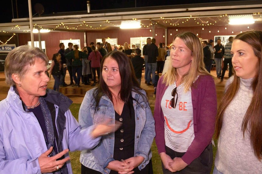 A woman wearing a mauve jacket stands talking next to three women outside a shed with fairy lights in the background.