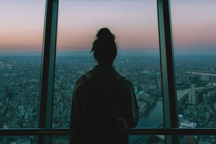 Colour photograph of a person looking out of a high rise building window at the Tokyo skyline.