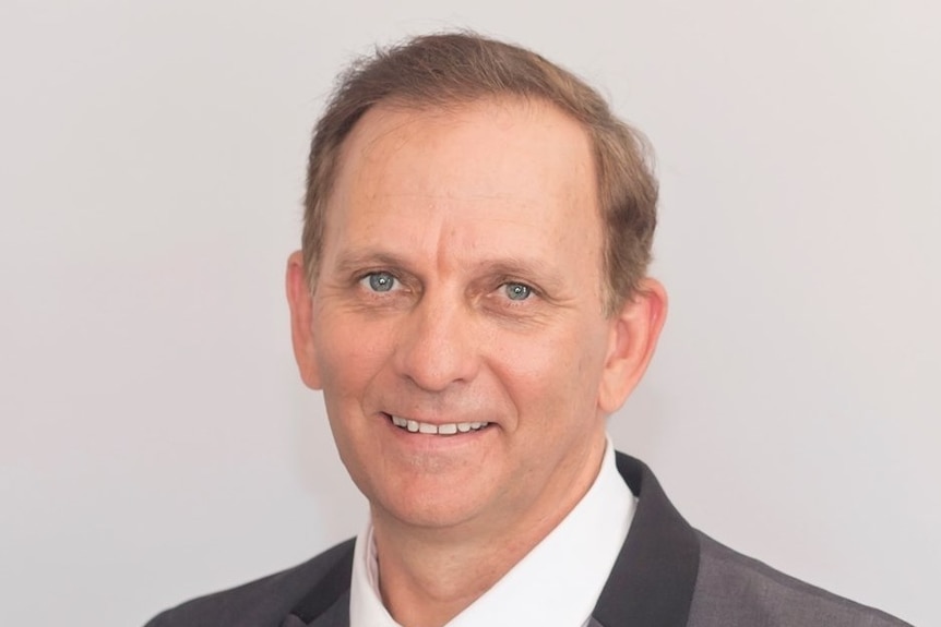 A man with brown hair smiles. He is wearing a dark grey suit, white shirt and grey and purple striped tie.