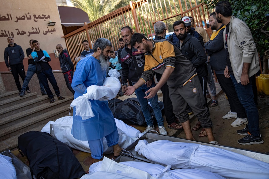arab men crowding around body bags as one doctor hands one man a smaller body bag