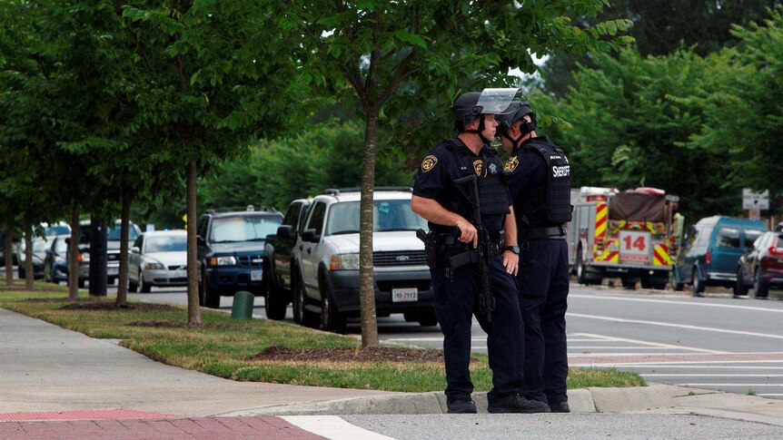 Two police officers look in opposite directions on, with the closest officer placing his hand on his gun. They stand in street.