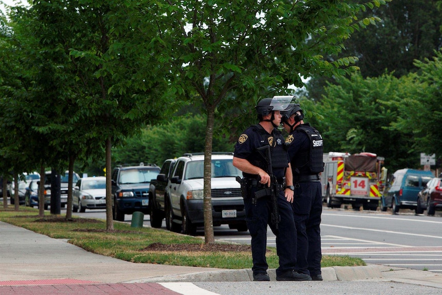 Two police officers look in opposite directions on, with the closest officer placing his hand on his gun. They stand in street.
