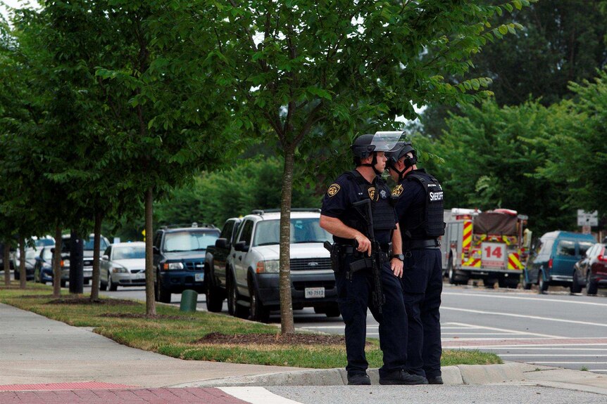 Two police officers look in opposite directions on, with the closest officer placing his hand on his gun. They stand in street.