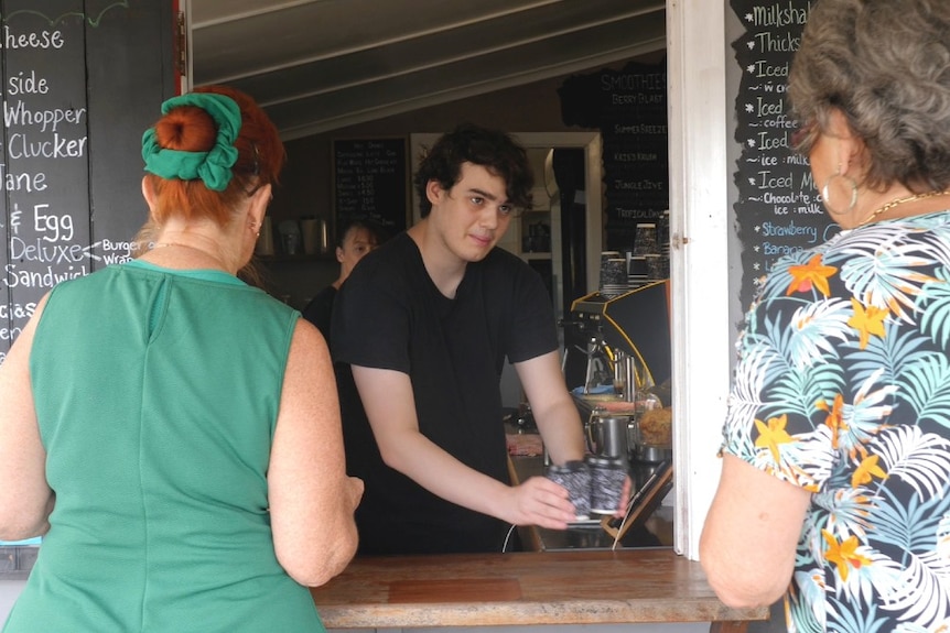 Young man, Isaac delivers two coffees to two women who have their backs to the camera.