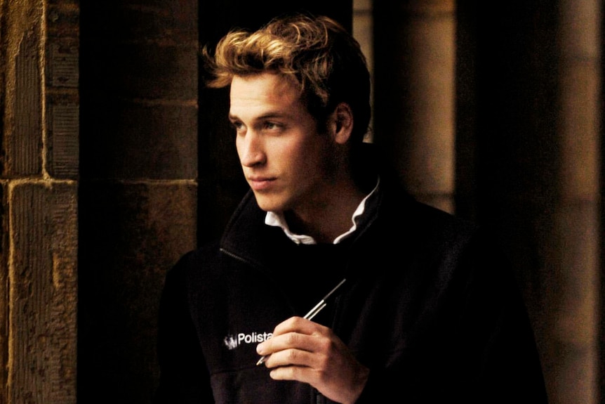 Le prince William adolescent à l'université, tenant un cahier et regardant ailleurs.