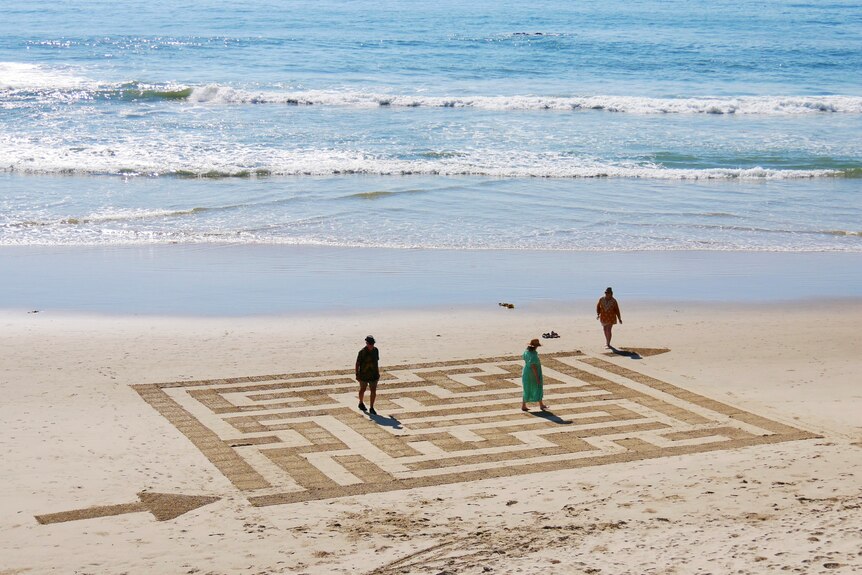 A maze design on the sand at low tide at a beach, with people walking through the maze pathways.
