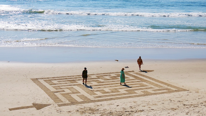 A maze design on the sand at low tide at a beach, with people walking through the maze pathways.