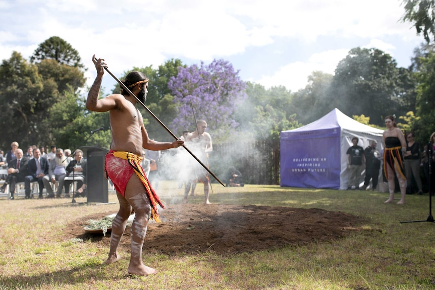 An Aboriginal male in traditional clothing aims a spear at the ground in front of onlookers as smoke rises from a dish
