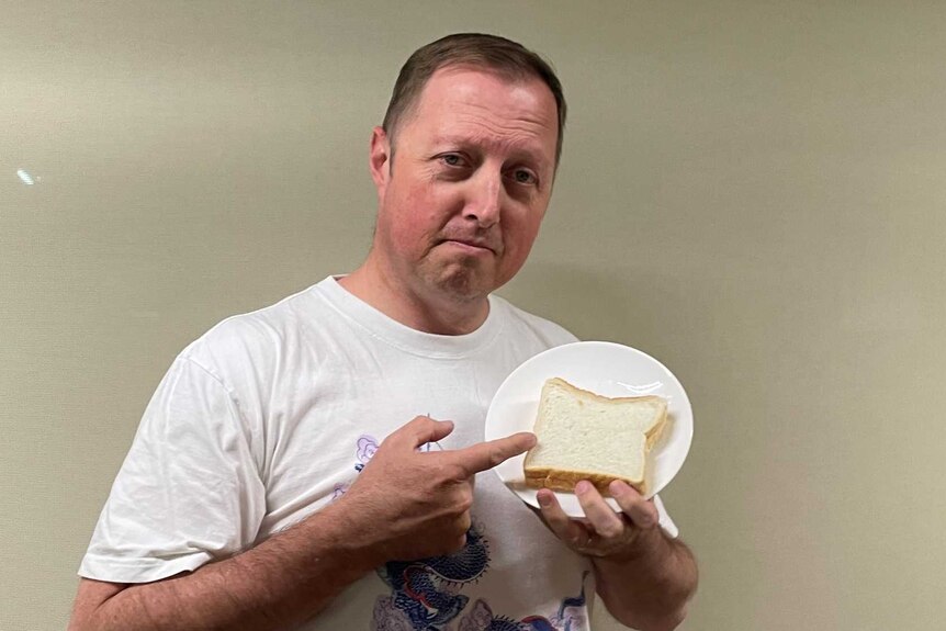 A man with short hair, wearing a white shirt, points solemnly at a sandwiich on a plate.