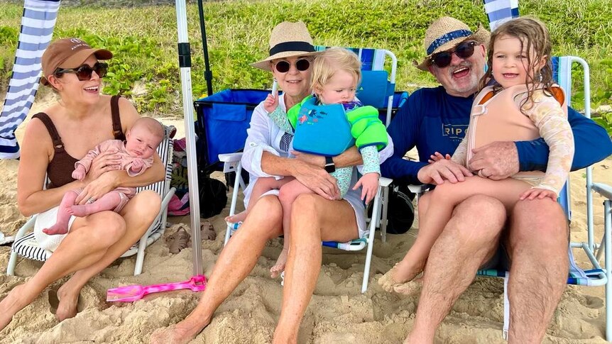 Three adults holding three young children lounging on beach chairs on the sand.