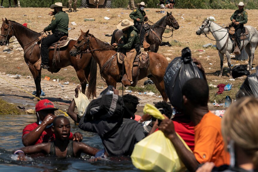 Migrants seeking asylum in the US cross a river as guards on horseback look on.