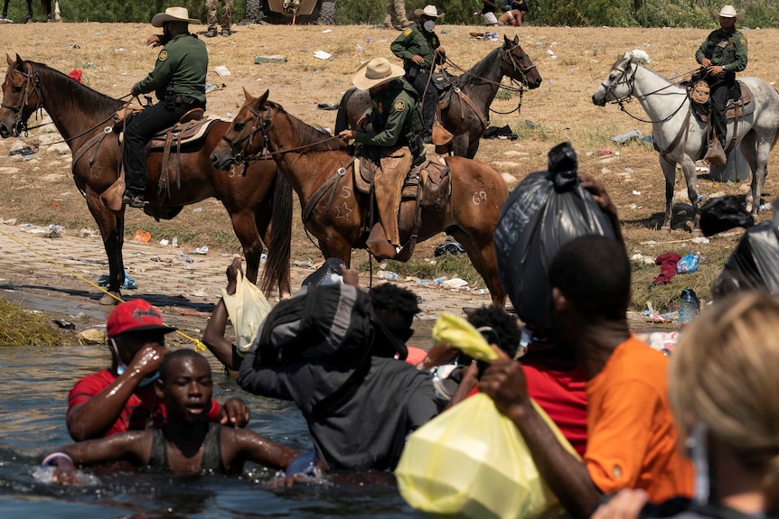 Migrants seeking asylum in the US cross a river as guards on horseback look on.