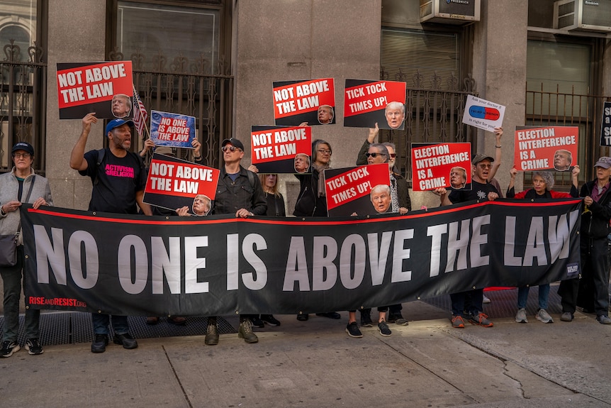 People hold a banner and signs that say 'no one is above the law' outside a courthouse building.