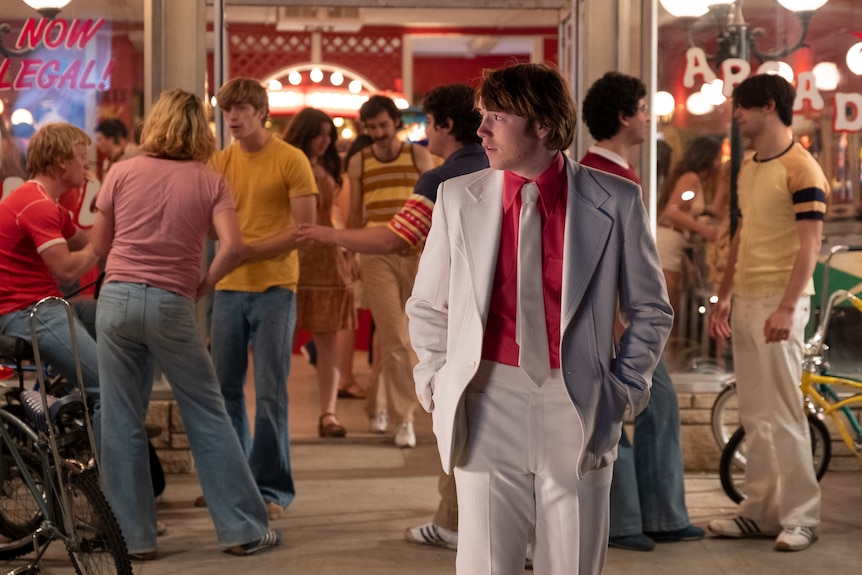 A brown-haired teenage boy in a red shirt and a white suit and tie stands in front of a crowded 70s arcade, looking expectant
