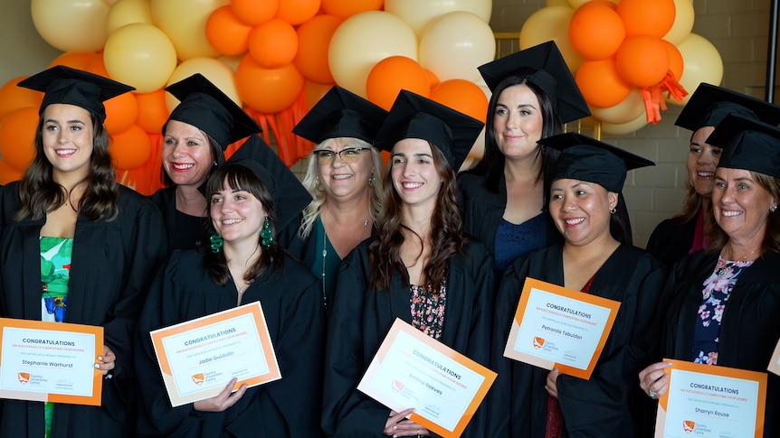 Nine women wearing black university graduation gowns holding certificates. 