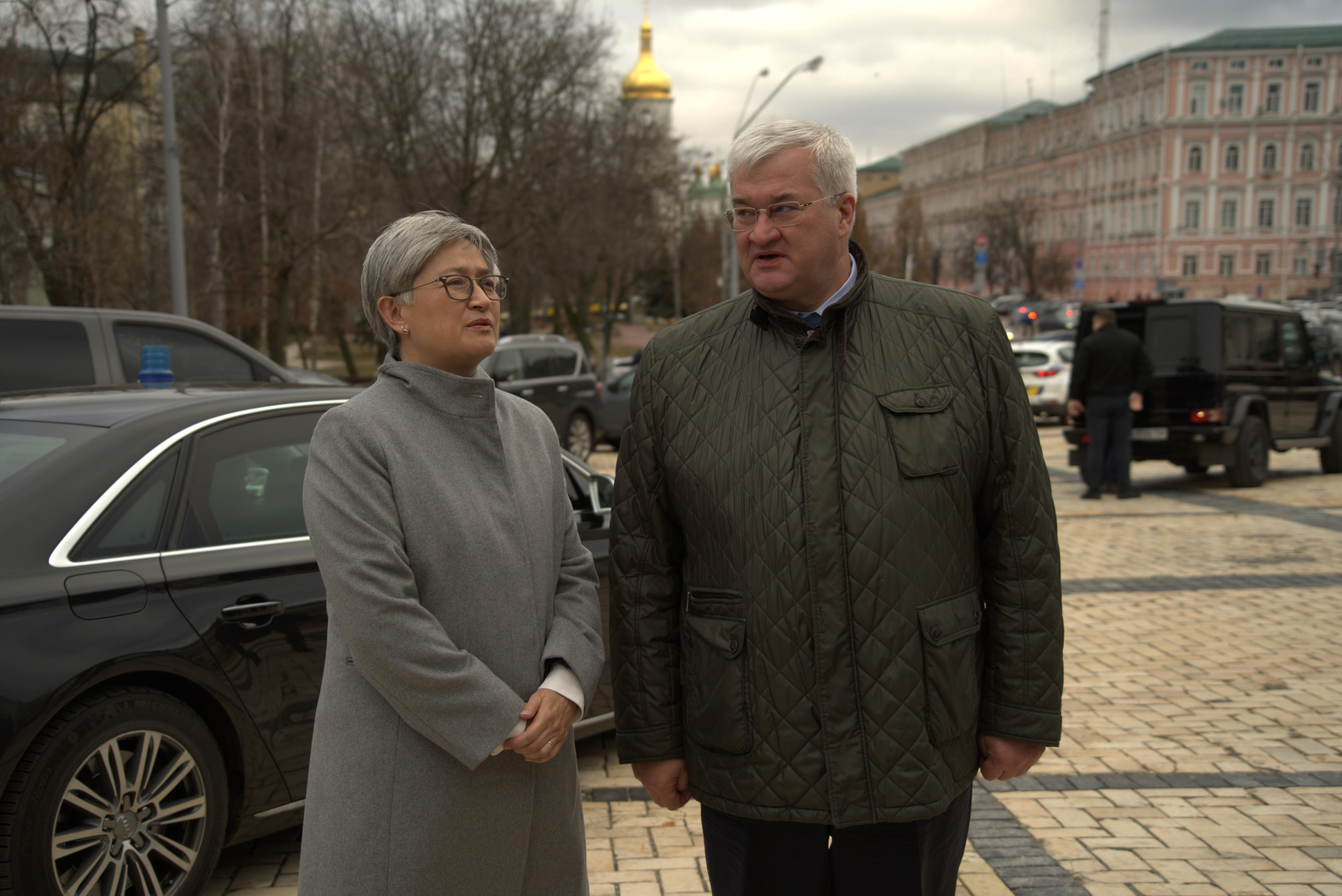 A woman in a grey coat stands next to a man in a green coat in front of a black car talking to each other