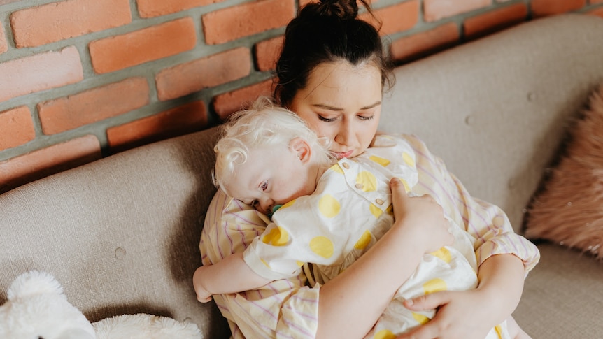 A woman with dark hair sits on a sofa cuddling a baby with blonde hair and chewing on a dummy