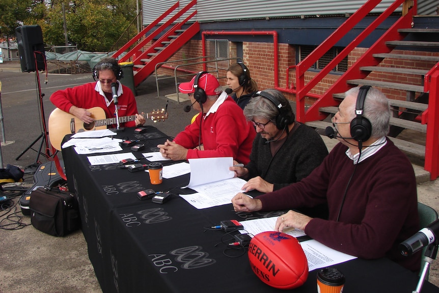 A table of three older men and one younger women, sitting outside a sports grandstand for a radio broadcast. One has a guitar