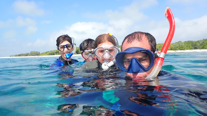 A man, woman and two children wearing scuba masks in the ocean with an island shore behind them