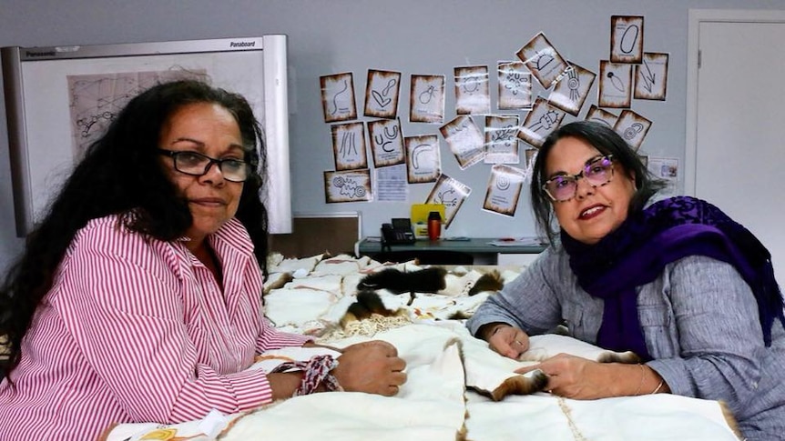 Two Aboriginal women working at a table sewing together squares of possum skin