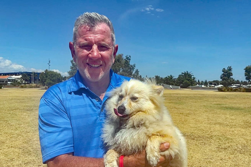 Older man wearing a blue shirt and holding a dog, standing on a field.