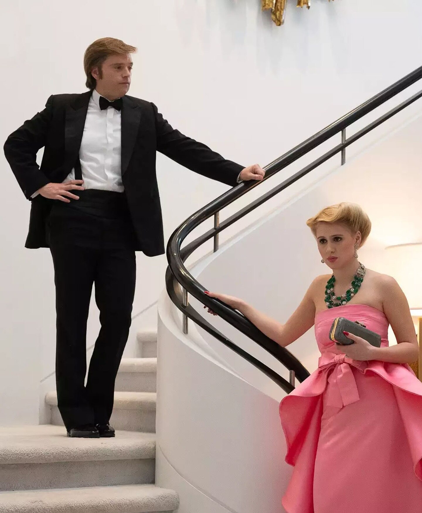 Trump stands impaitently on a curved, white carpeted staircase. His wife Ivana stands below in a pink dress