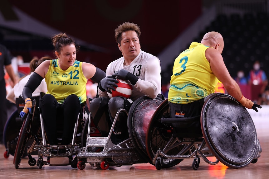 A woman in a wheelchair wearing a green and gold singlet pushes to reach a male opponent holding the ball