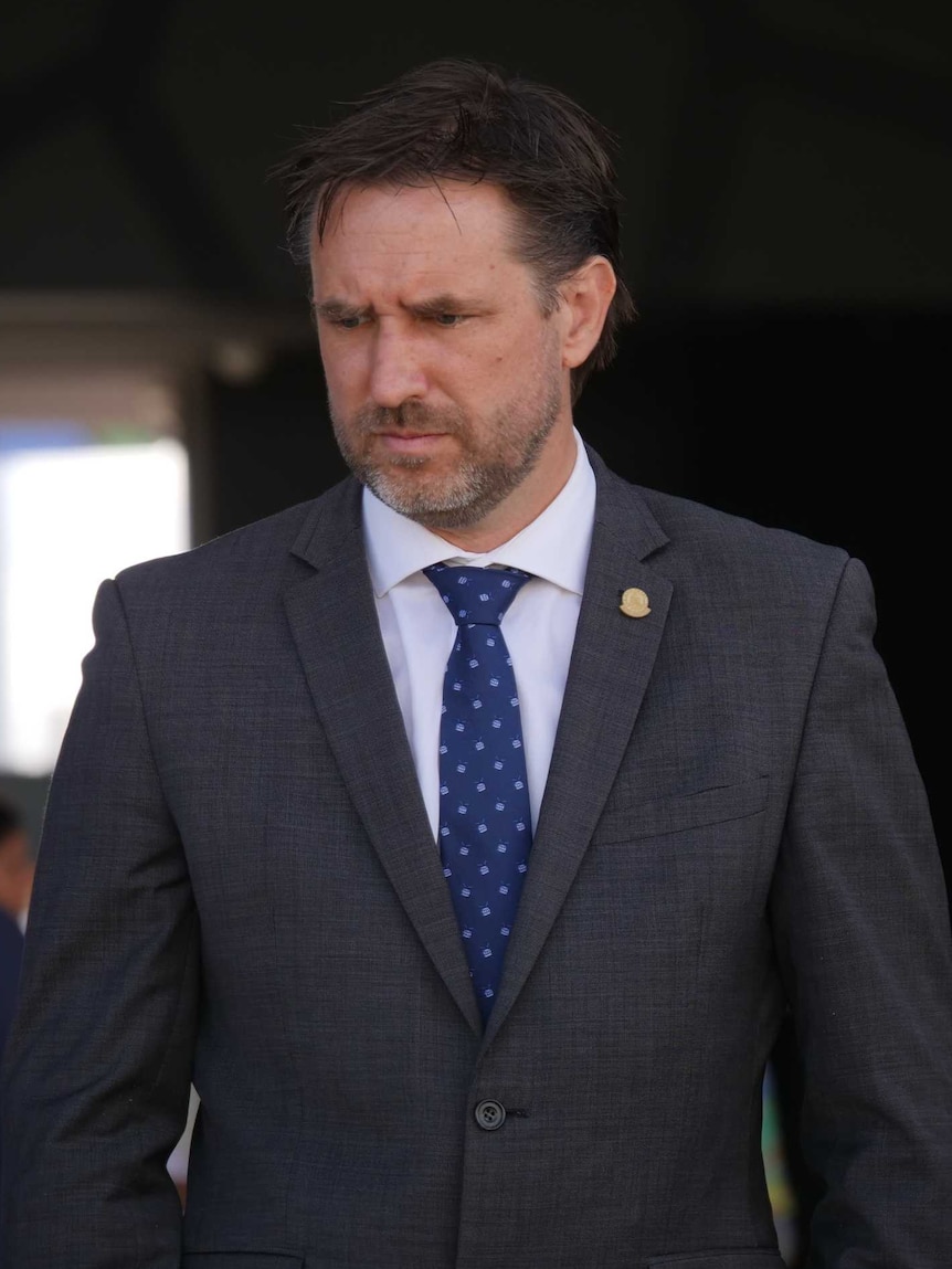 A aman in a grey suit and blue tie looks away from the camera