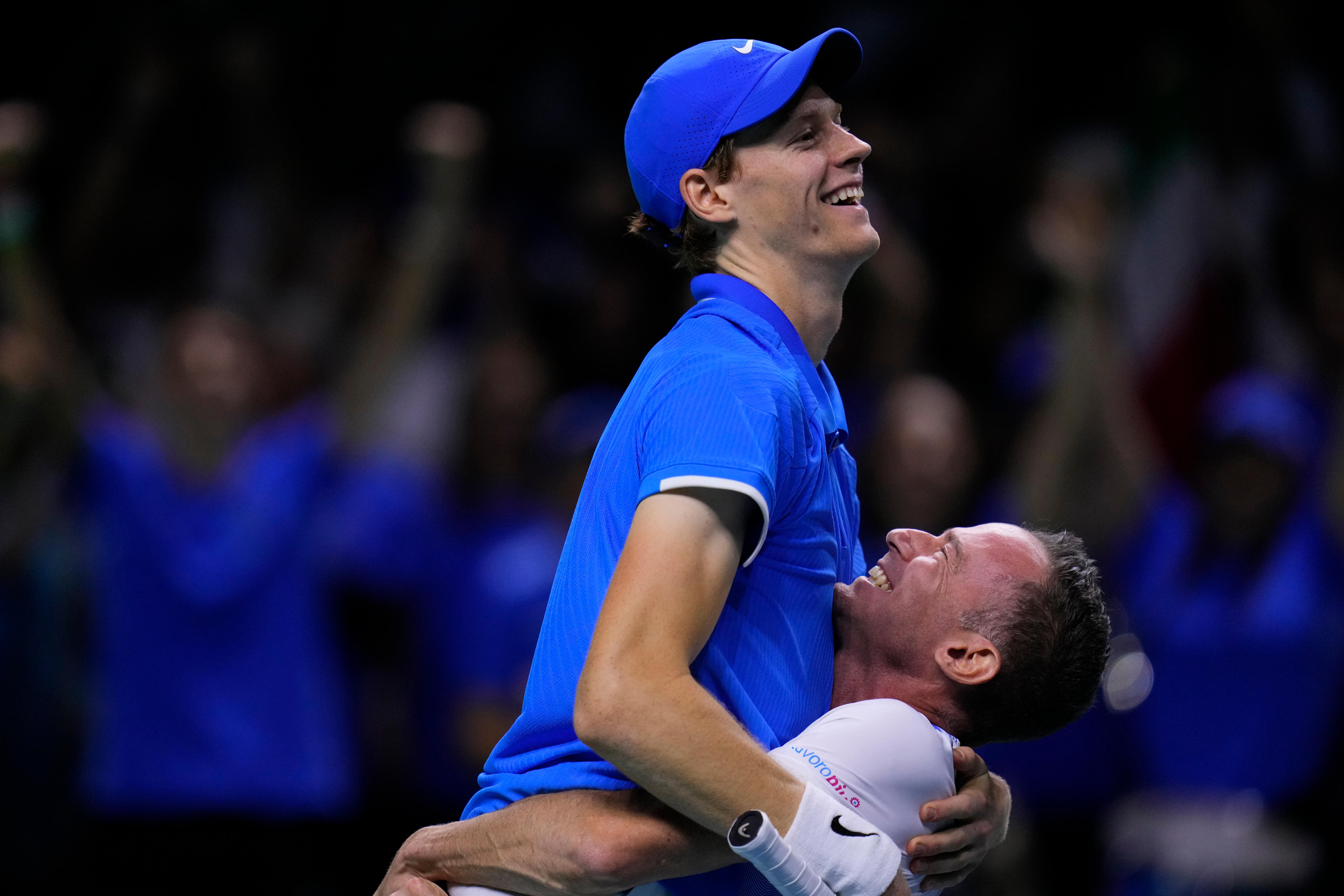 An Italian Davis Cup player wearing a blue cap grins as he is lifted in the air in celebration by his team captain.