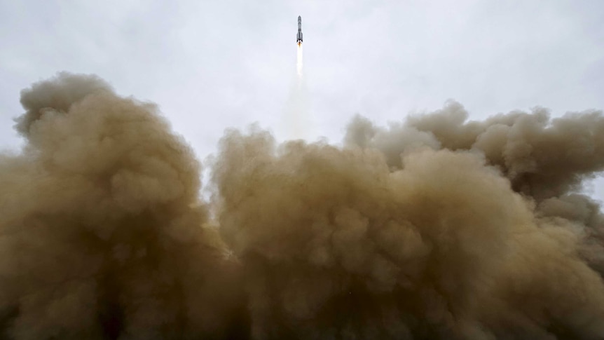 The Proton-M rocket, small in frame, appears over the smoke and dust from the blast off, large in the foreground.