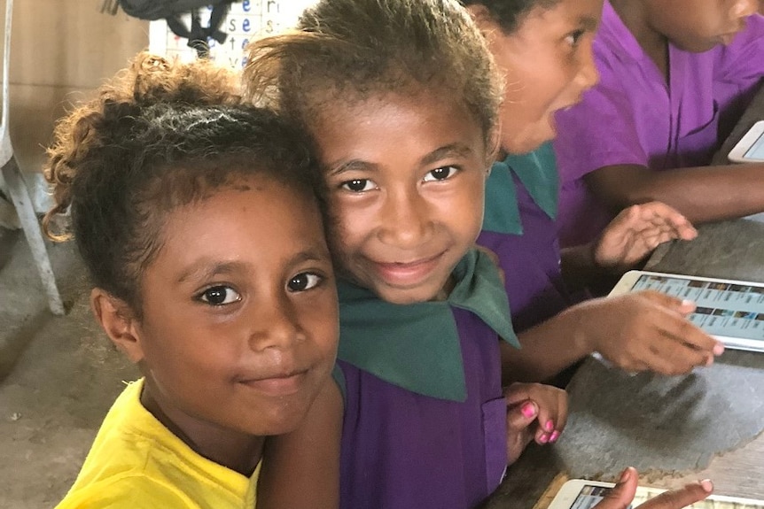 Two young school girls sit at a desk smiling, reading from a tablet.