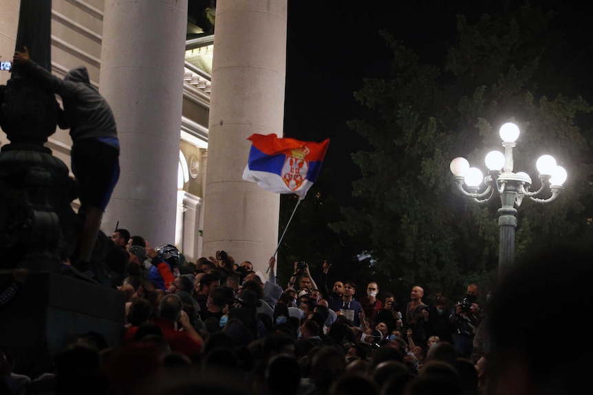 A mob gathers waiving a single flag on the steps of a government building with large white marble columns.