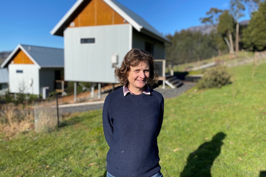 A woman stands in front of a small cabin on a property. There is green grass and blue sky in the background